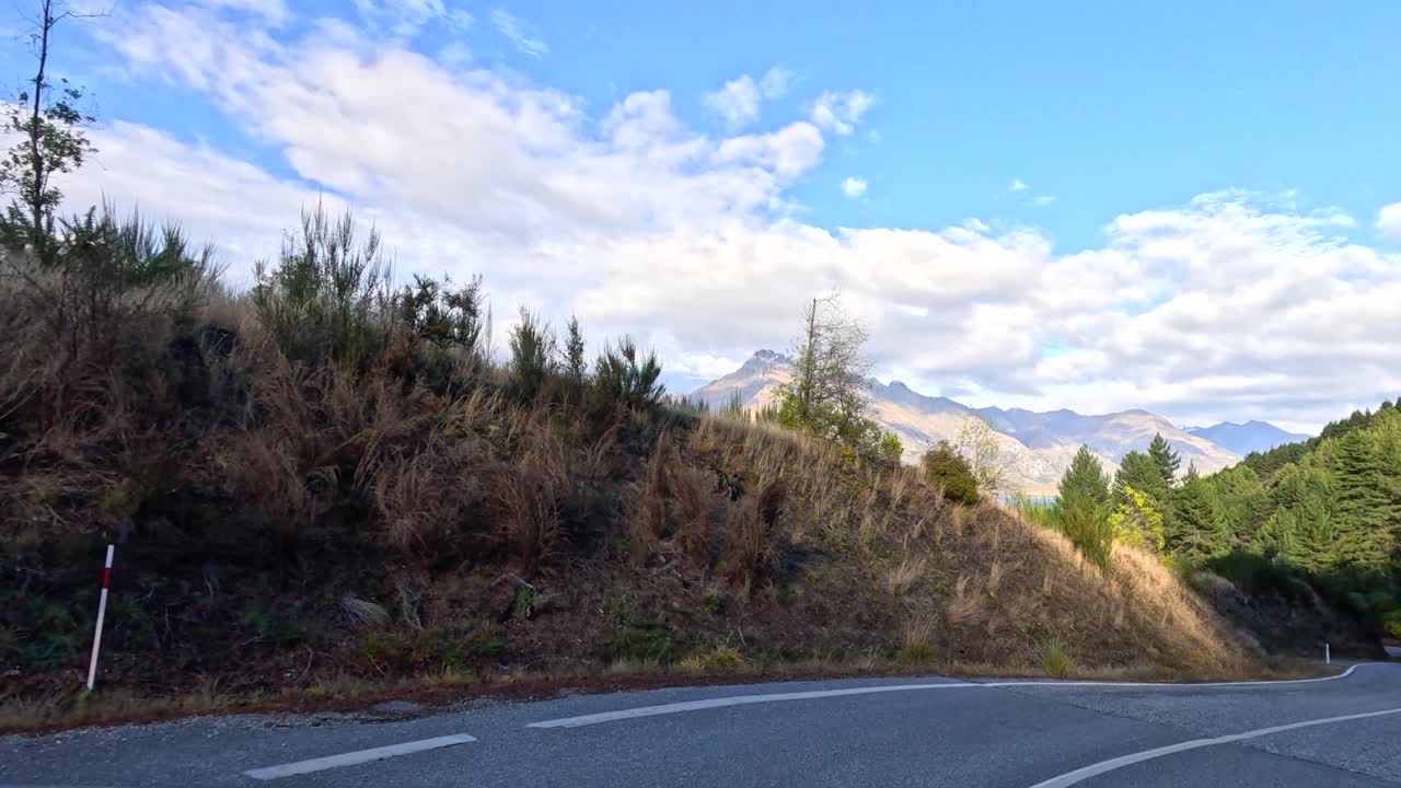 Vehicle travels winding mountain road, surrounded by trees, mountains, and bright daylight in Queenstown