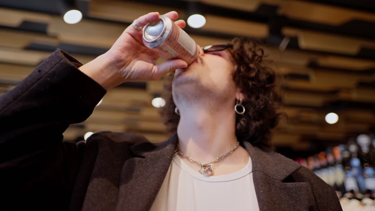 un hombre moreno confiado y fresco con cabello rizado y gafas de sol negras camina a través de un supermercado y bebe limonada durante sus compras