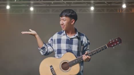 adolescente asiático con guitarra sonriendo y señalando al lado en el escenario