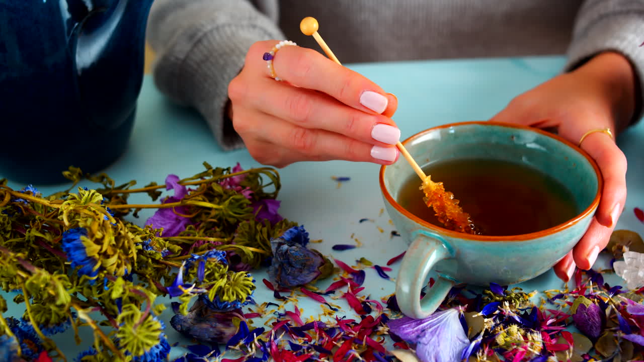 Woman stirring and drinking warm tea with brown sugar candied sticks. Cup of steaming hot tea on light blue background with flowers, leaves and candy sticks. Slow motion of woman drinking herbal tea.