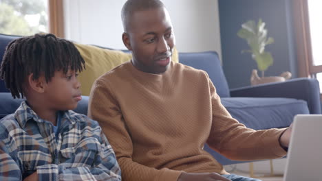 Happy african american father and son sitting on floor and using laptop at home, slow motion