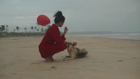 toma cinematográfica de una modelo de moda india con un vestido rojo y llevando un globo rojo acariciando a un perro salvaje en goa india en una playa de arena en un día ventoso