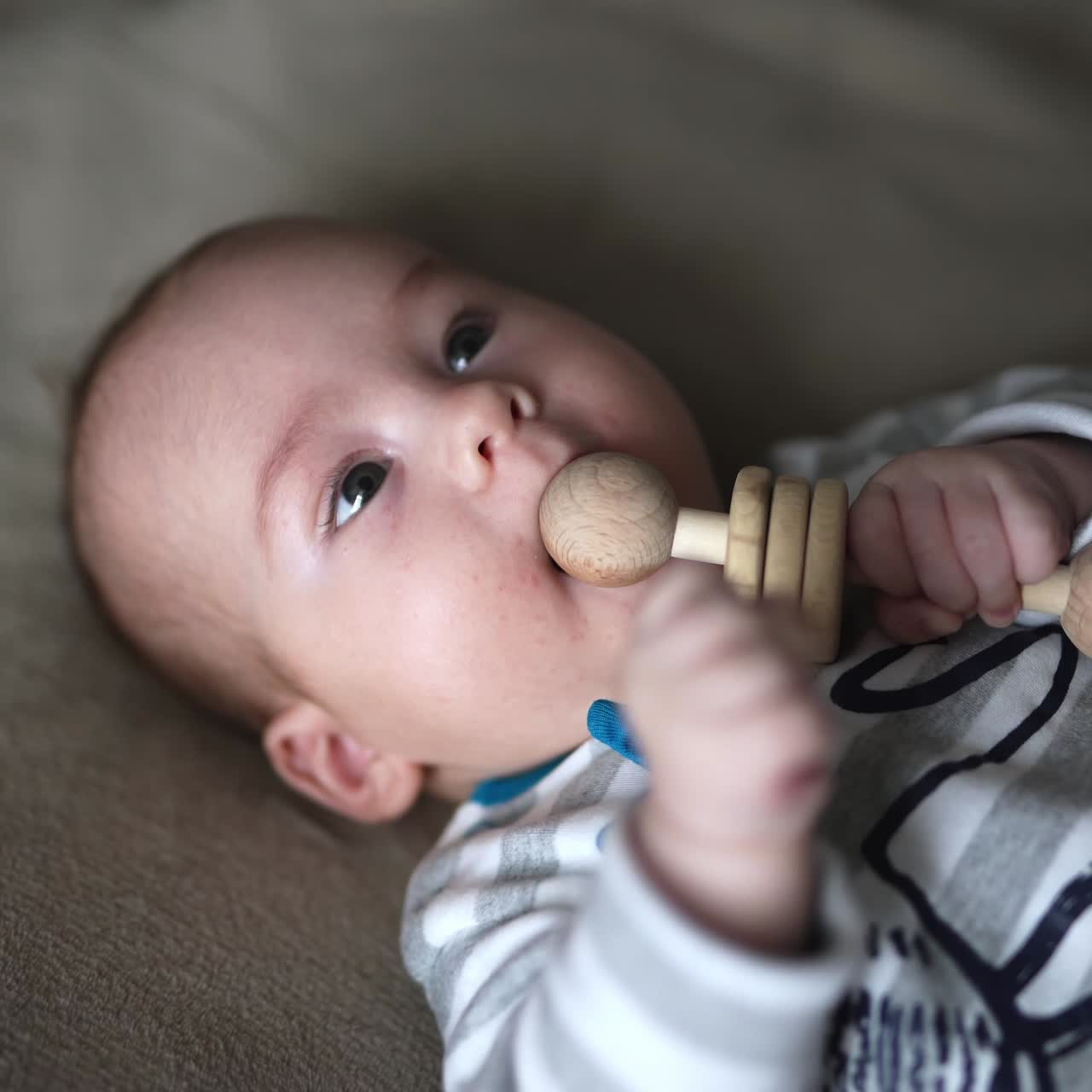 Cute kid on the bed lies his face up. Beautiful baby holding wooden toy in his hand and tries to bite it. Close up