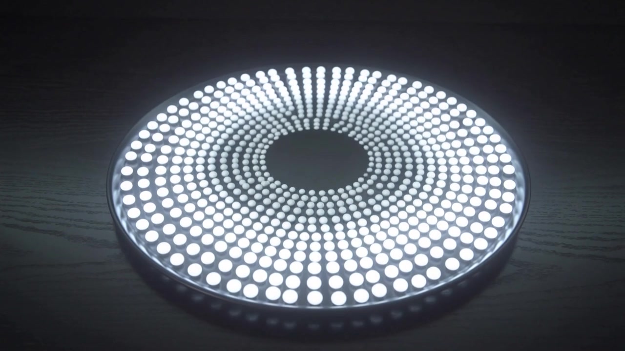 Panel LED circular encendiéndose, formando anillos concéntricos brillantes hacia afuera sobre una mesa de madera oscura, difusor.