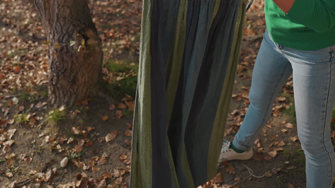 Woman folding hammock on forest floor, wearing green sweater and jeans, hands smoothing striped fabric among fallen leaves, prepping hammock for storage and transport, calm autumn light, peaceful