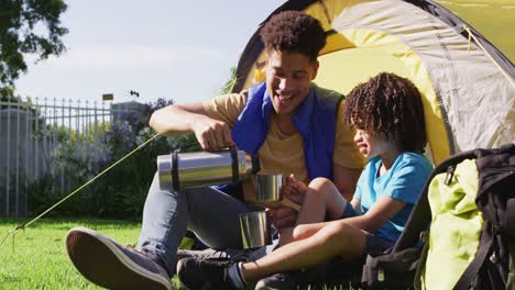 Happy-biracial-man-and-his-son-drinking-from-thermos-flask-in-garden