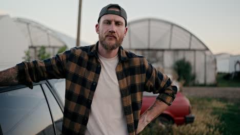 portrait of a serious farmer guy with a beard in a checkered shirt posing against of greenhouses on the farm