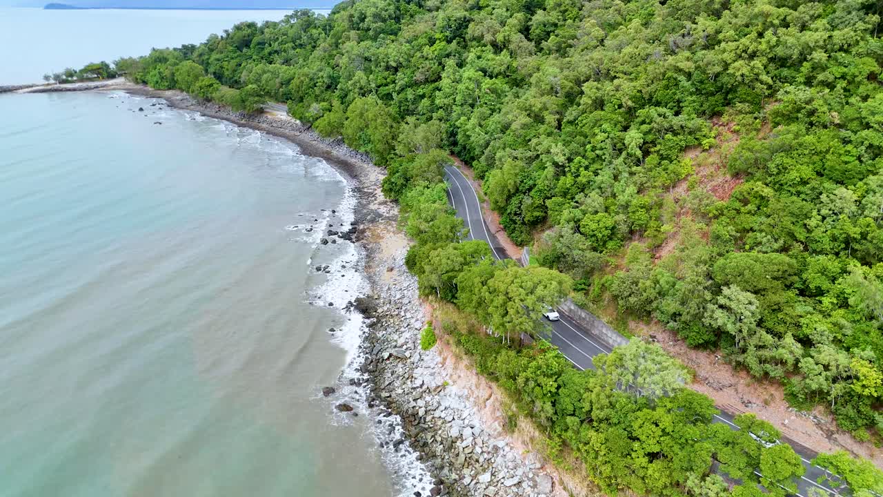 Drone follows car along winding coastal road beside lush rainforest and ocean, daylight, smooth movement
