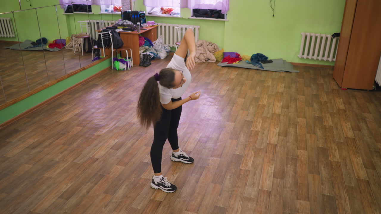 Dáma ve sportovním oblečení se protahuje uvnitř, zvedá jednu ruku vzhůru a ohýbá se ze strany na stranu na dřevěné podlaze, udržuje rovnováhu a držení těla ve fitness studiu se zrcadlem, židlí, šatní skříní a dalšími předměty