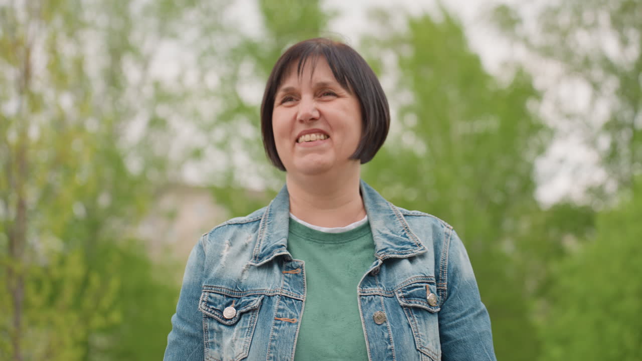 Caucasian Woman Looking Up Smiling Amid Spring Foliage In Park Wearing Denim Jacket And Green Shirt, Candid Frontal Portrait With Hopeful Gaze And Natural Light, Gestures Suggest Speaker, Mentor