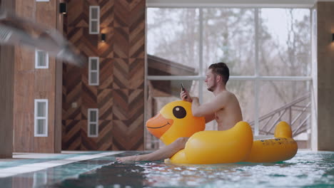 un hombre adulto está usando un teléfono móvil mientras descansa en la piscina sentado en un pato inflable y flotando en la piscina