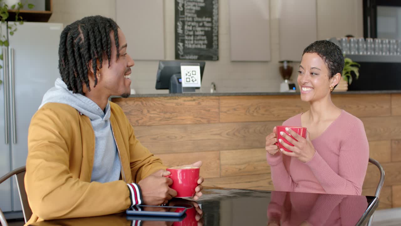 Diverse couple clinking red mugs sharing talk over coffee across cafe table beside POS terminal