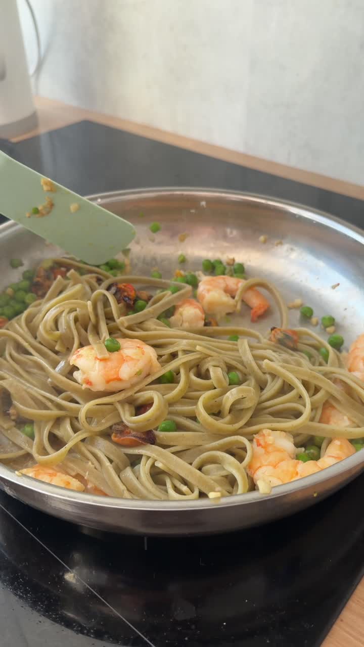 Mano che mescola pasta verde con gamberetti e piselli in padella