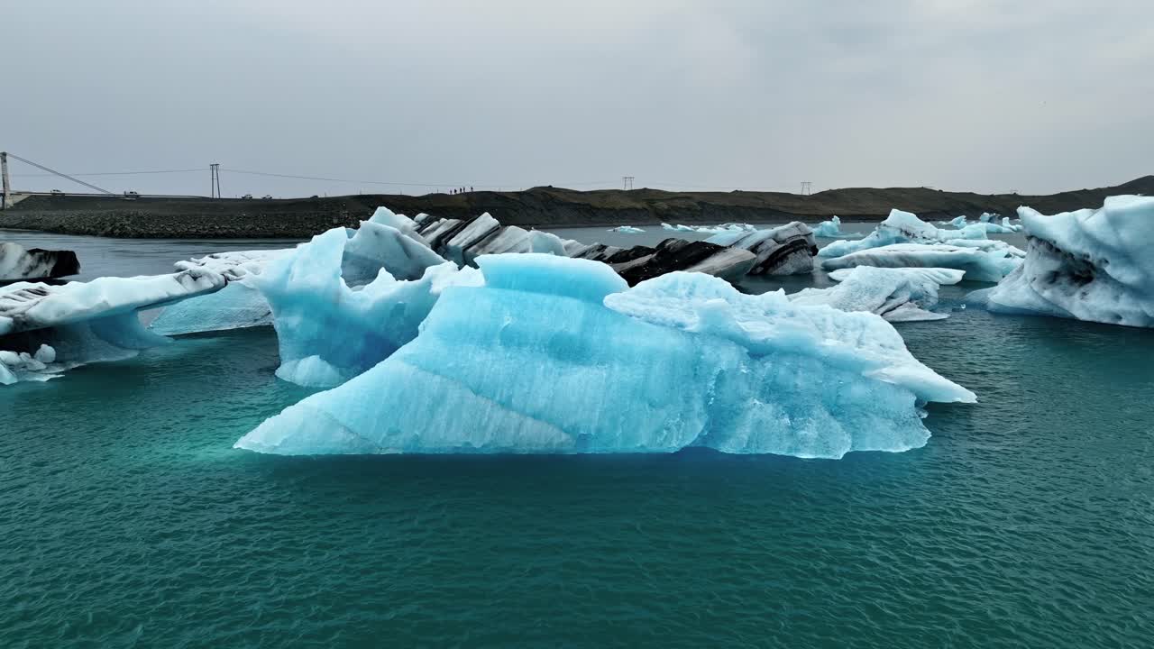 Calm drone sweep over striking blue icebergs drifting in cold Icelandic water, framed by a distant bridge and rugged shoreline, highlighting dramatic shapes, layered ice tones, and Arctic atmosphere