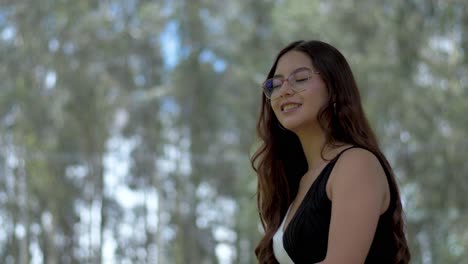 Elegant-Hispanic-woman-with-open-hair-and-glasses-enjoys-a-cheerful-conversation-in-nature's-embrace,-surrounded-by-towering-trees