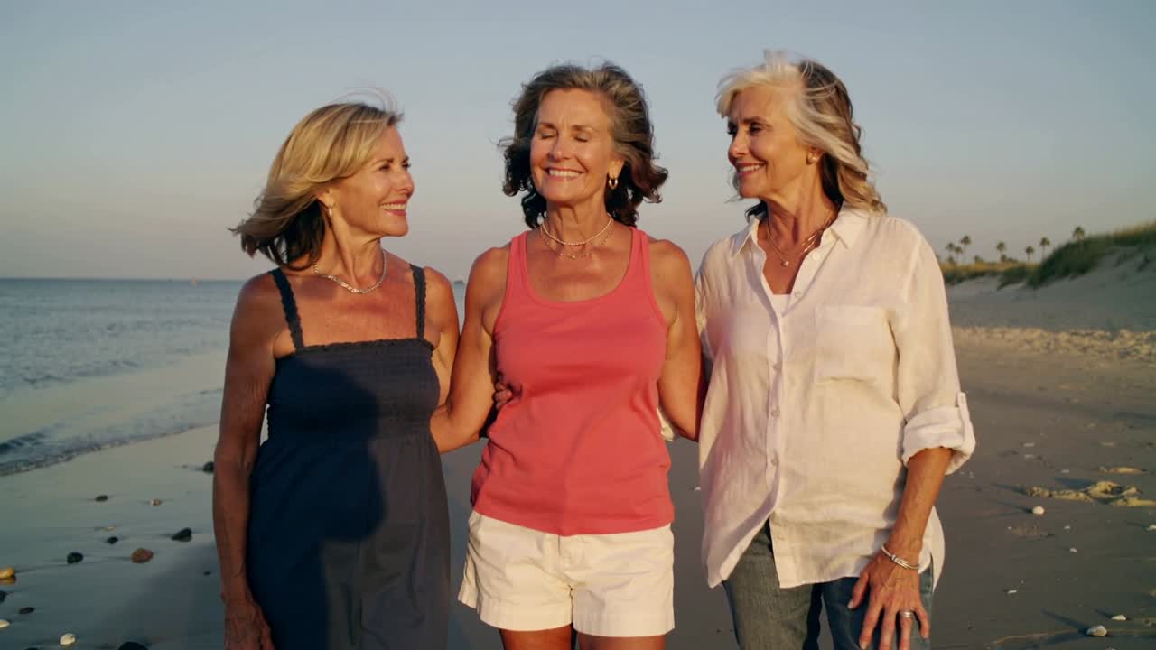 Walking senior trio linking arms on beach, breeze lifting hair, in coral, navy, white as friends