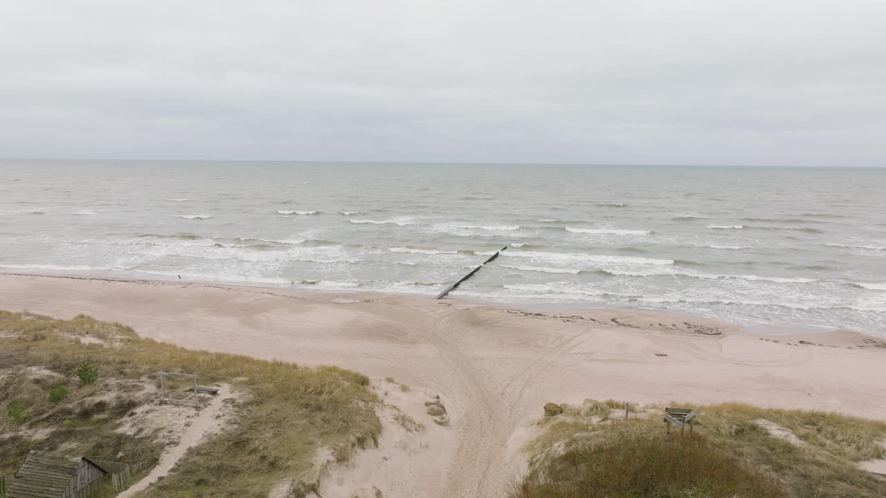 съемка с воздуха побережья балтийского моря в пасмурный день, старый деревянный пирс, белый песчаный пляж, большие штормовые волны бьются о берег, изменения климата, широкая съемка с дрона, движущаяся вперед