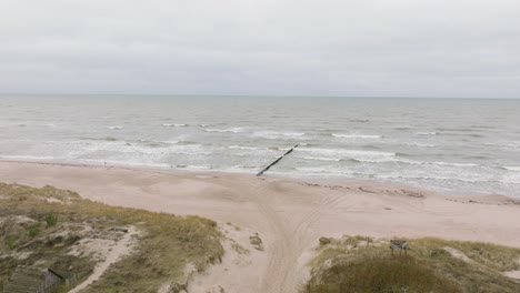 establishing aerial view of baltic sea coast on a overcast day, old wooden pier, white sand beach, large storm waves crushing against the coast, climate changes, wide drone shot moving forward