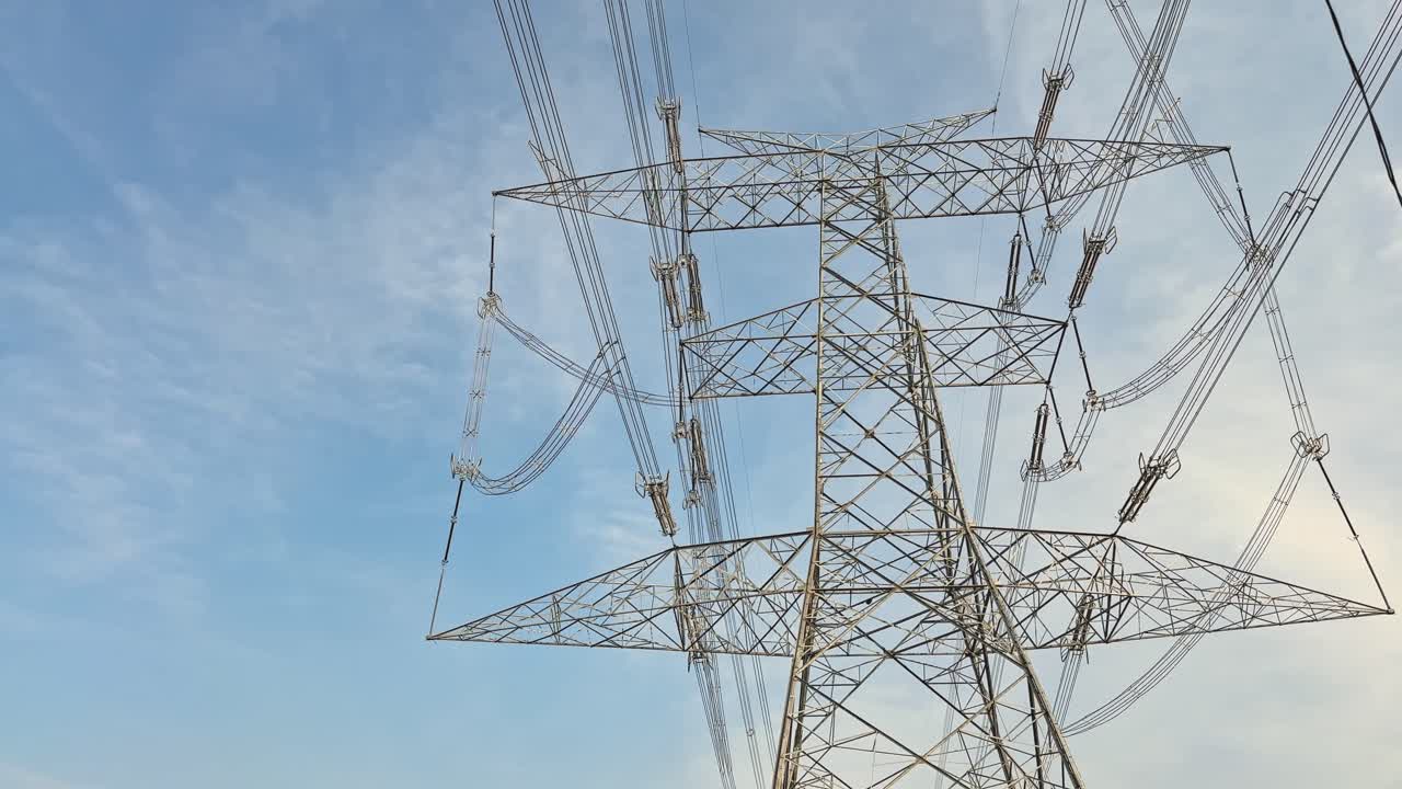 Slow pan across a high-voltage transmission tower, steel lattice arms holding bundled conductors and insulators, power lines sweeping against clear blue sky in daylight slowly panning shot up