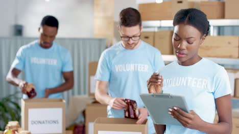 joven voluntaria afroamericana escribiendo y revisando donaciones en un almacén de caridad mientras sus compañeros de trabajo clasifican cosas donadas en cajas en el fondo