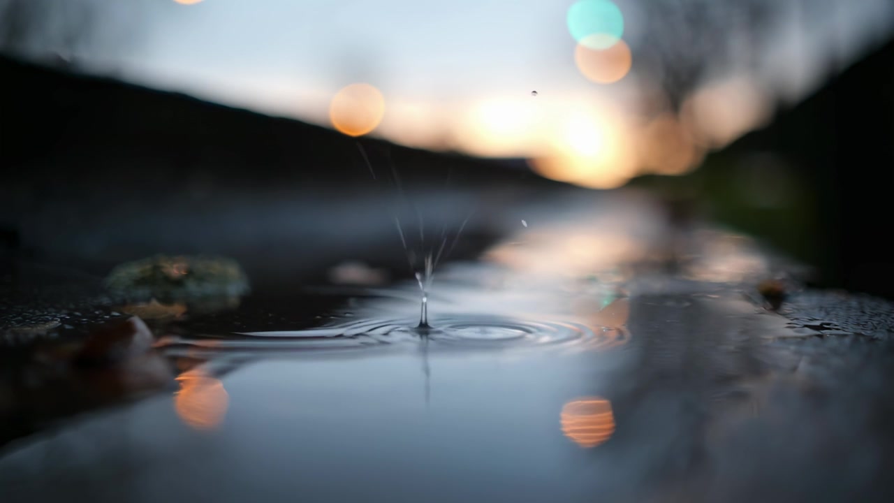 ボケライトを背景に、水たまりに波紋を作る雨粒のクローズアップ