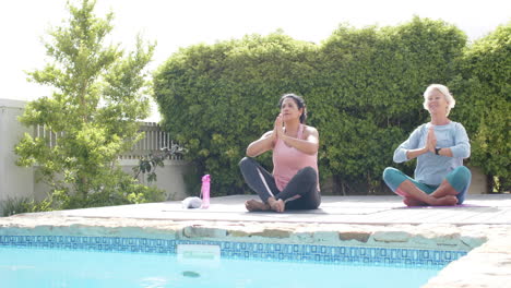 dos mujeres mayores felices y diversas practicando yoga junto a la piscina en un jardín soleado, cámara lenta, espacio de copia