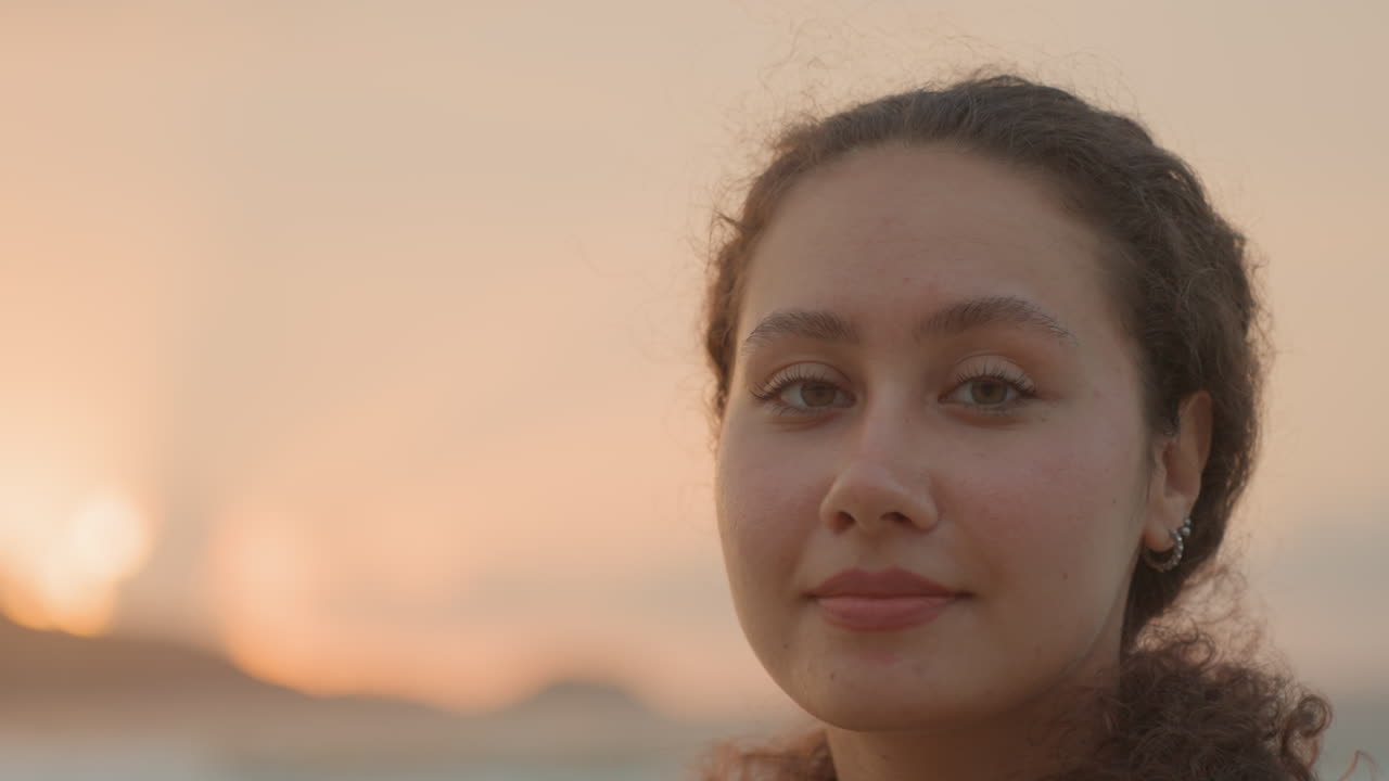 Young woman island shore sunset smiling camera, warm golden hour glow, direct gaze, playful expression, curly hair, earring detail, gentle waves, closeup portrait, influencer content mood, relaxed