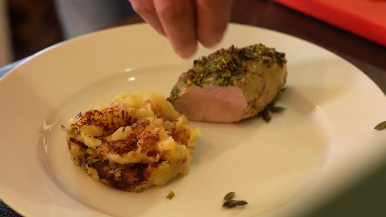 Person carefully places slice of meat topped with herbs on plate, potato side dish.