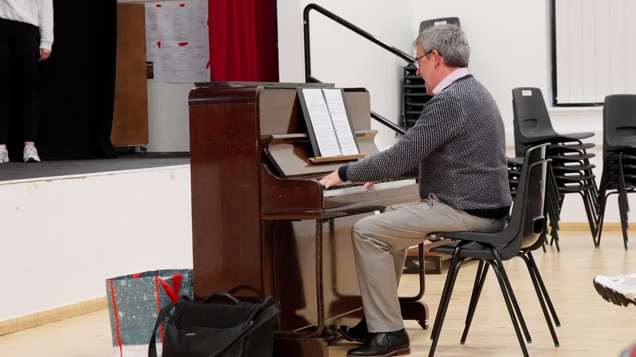 Man plays piano during indoor rehearsal in community hall, surrounded by chairs and music sheets. Captured in venue lighting