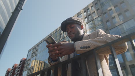 Black-Businessman-Leaning-on-Railing-and-Typing-on-Smartphone