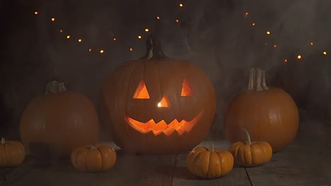 Static-shot-of-candle-lit-Jack-o-lantern-in-fog-on-a-wooden-surface-with-a-dark-background-and-orange-lights