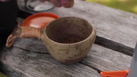 persons hands making instant camping coffee in a wooden kup with leather handle on a bench