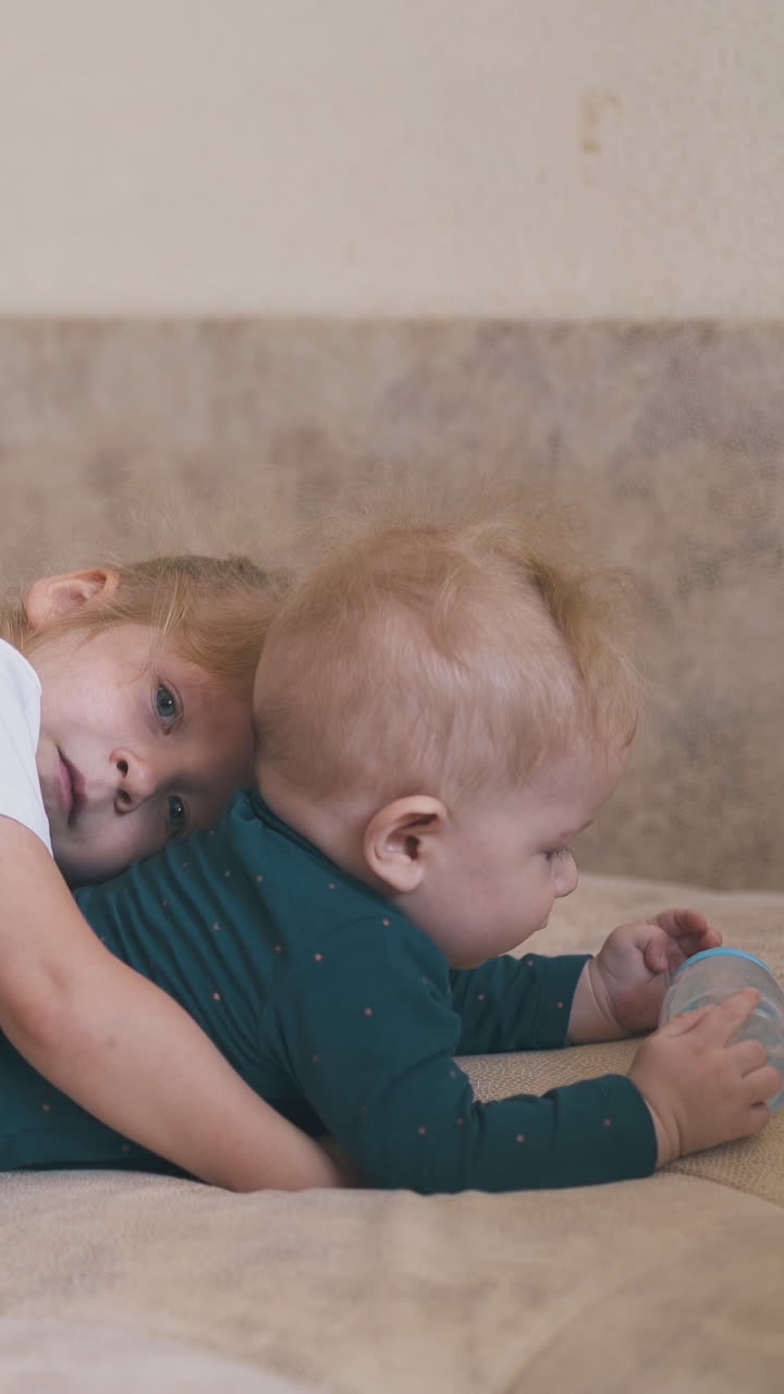 cute blonde girl hugs funny baby brother with bottle of water lying on large bed in spacious room at home