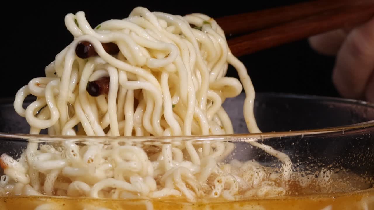 黒い背景を背に、木製の箸がガラスの丼から湯気の立つインスタント麺を持ち上げています。