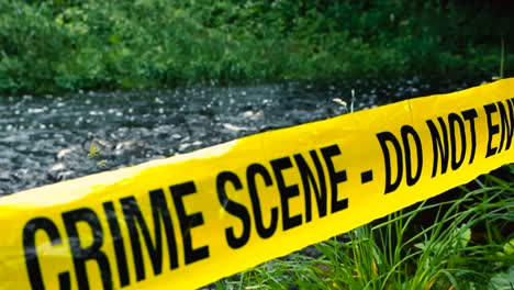 yellow crime scene do not enter police ribbon tape pulled in front of a foamy and fast flowing river in the countryside during summer cloudy day. shoreline is grassy and water is dark with white water