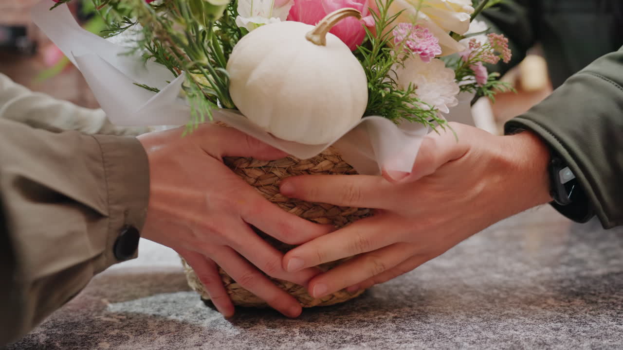 El mostrador de una floristería recibe un ramo tejido con calabaza blanca y flores rosadas. Las manos colocan suavemente la cesta sobre una mesa de piedra texturizada. Papel de regalo rústico y cinta visibles en un interior cálido y vintage.