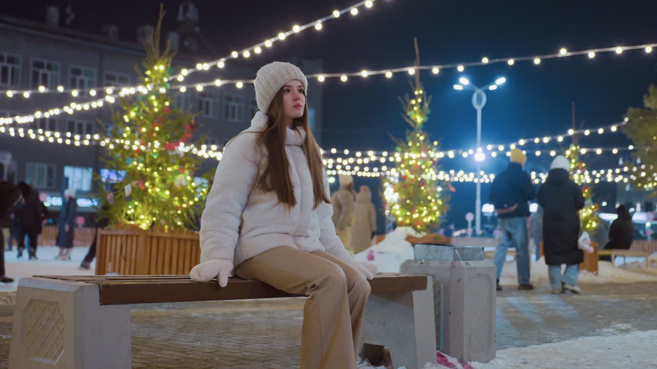 Woman in stylish winter coat and beanie walks towards wooden bench in vibrant city at night, festive lights glow in background as people skate and socialize