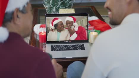 Padre-E-Hijo-Birraciales-Sonrientes-Usando-Una-Computadora-Portátil-Para-Una-Videollamada-Navideña-Con-Una-Pareja-En-La-Pantalla