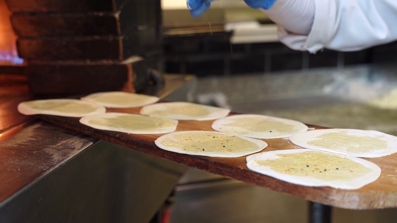 A gloved chef prepares traditional flatbread in a commercial kitchen. Close-up cinematic shot shows brushing dough with oil and sprinkling seeds on a wooden paddle before baking in a wood-fired oven.