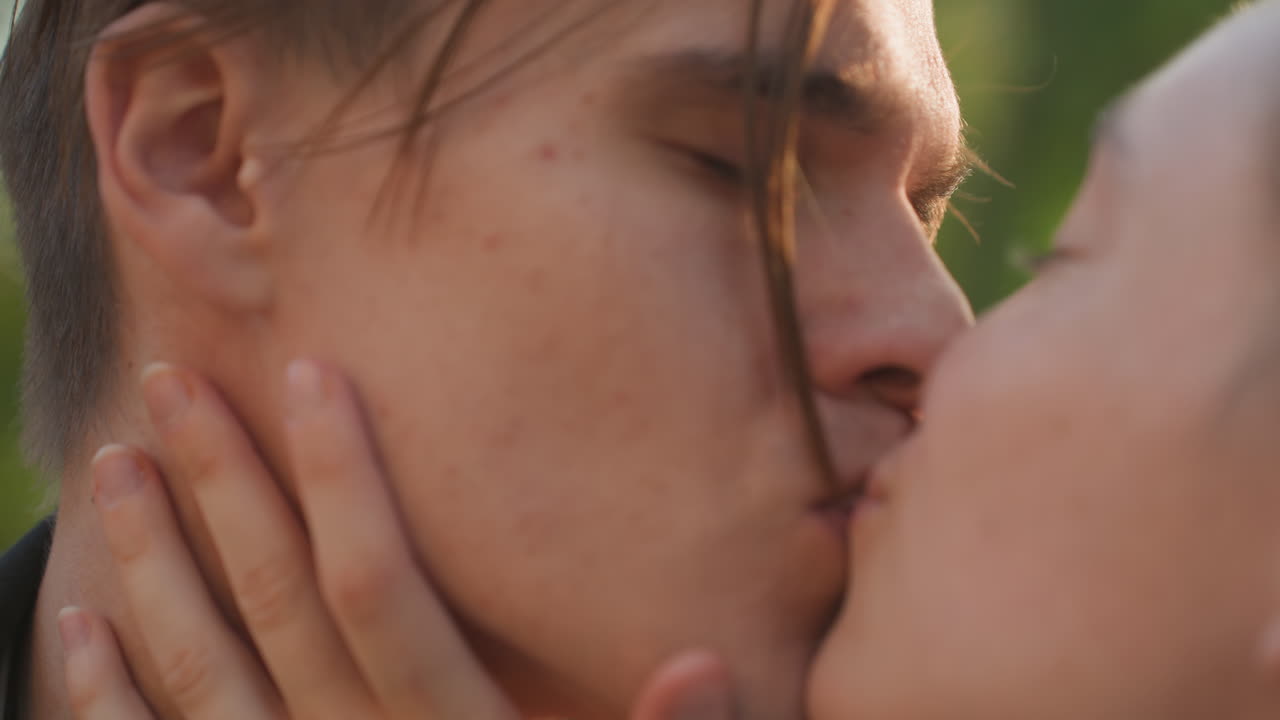 Primo piano di una coppia che si scambia un tenero bacio all'aperto, mani sul viso, morbida luce solare che filtra tra gli alberi, uno scambio romantico spontaneo che trasmette amore e impegno, dolce carezza e sorrisi affettuosi.