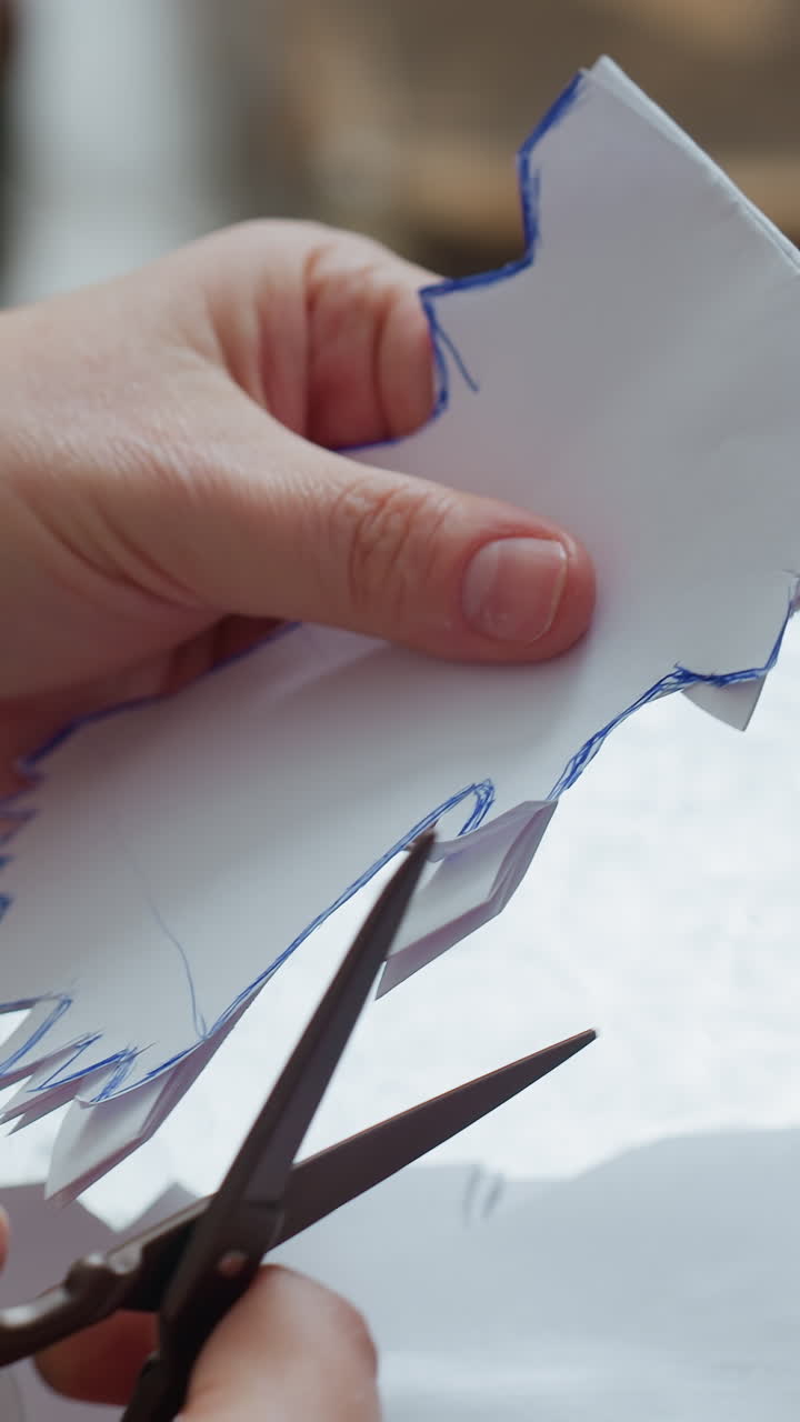 Primer plano de unas manos recortando un árbol de papel; escena de manualidades navideñas con tijeras recortando una plantilla impresa, luz natural suave y dedos concentrados guiando cortes precisos a través de hojas dobladas sobre una mesa