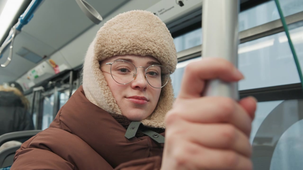 Nært portrett av ung kvinne sittende inne i et tog, iført beige sherpafôret trapperlue, klare briller og en brun isolert dunjakke, som støtter seg til en metallstang under en vintertur.