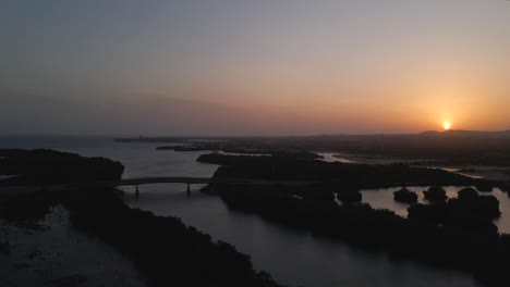 Drone-moves-to-the-right-gaining-altitude-revealing-a-silhouetted-bridges,-mangrove-forests,-ocean-on-the-horizon-and-mountains-with-the-majestic-sunset,-Porlamar,-Margarita-Island,-Venezuela