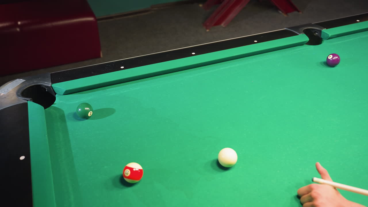 Up-close shot of hand using cue stick to strike white billiard ball on green pool table. Focus on scattered billiard balls near pocket, capturing motion and intensity of competitive gameplay