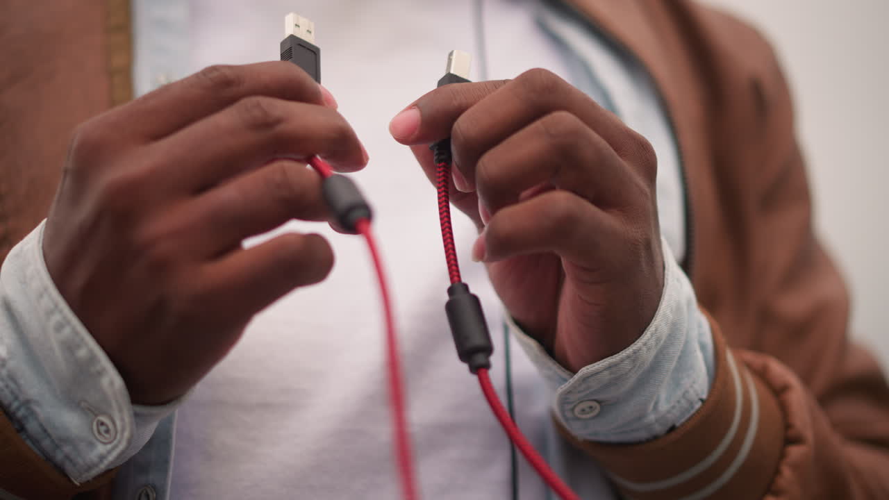 black DJ hands connecting red cable during street setup, closeup of connector and braided lead, leather jacket and denim cuffs, focused fingerwork preparing controller for urban night set