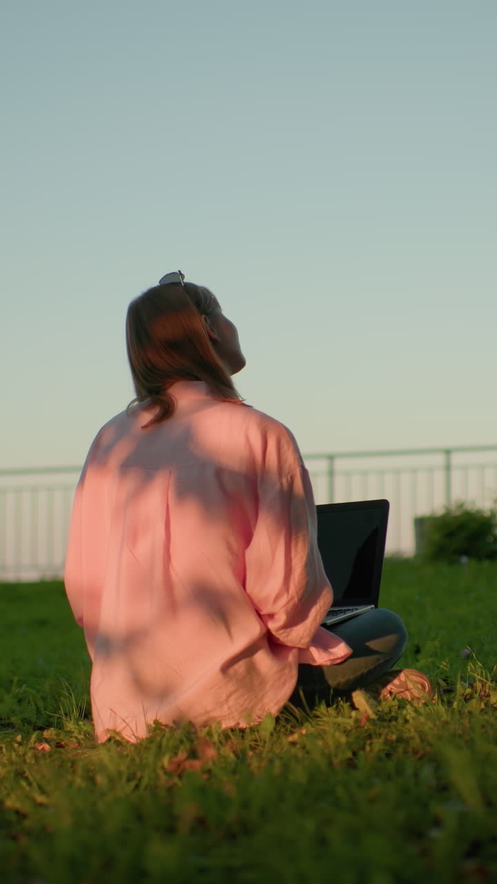 Back view of freelancer in pink top with glasses resting on her head looking up, focusing back on laptop, sunlight casting shadows on her clothes, bench and iron railing in distant background