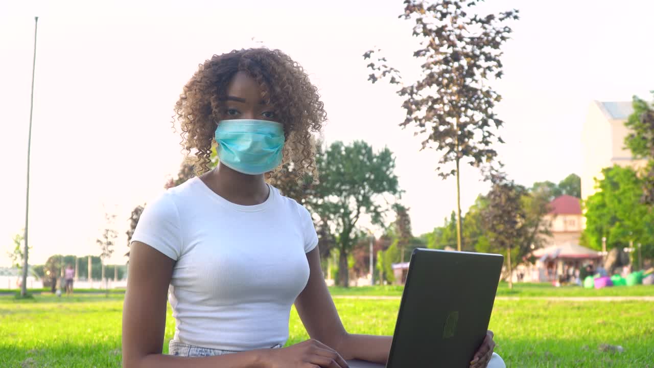 Young african american female student in protective mask working on laptop in park during covid 19 pandemic