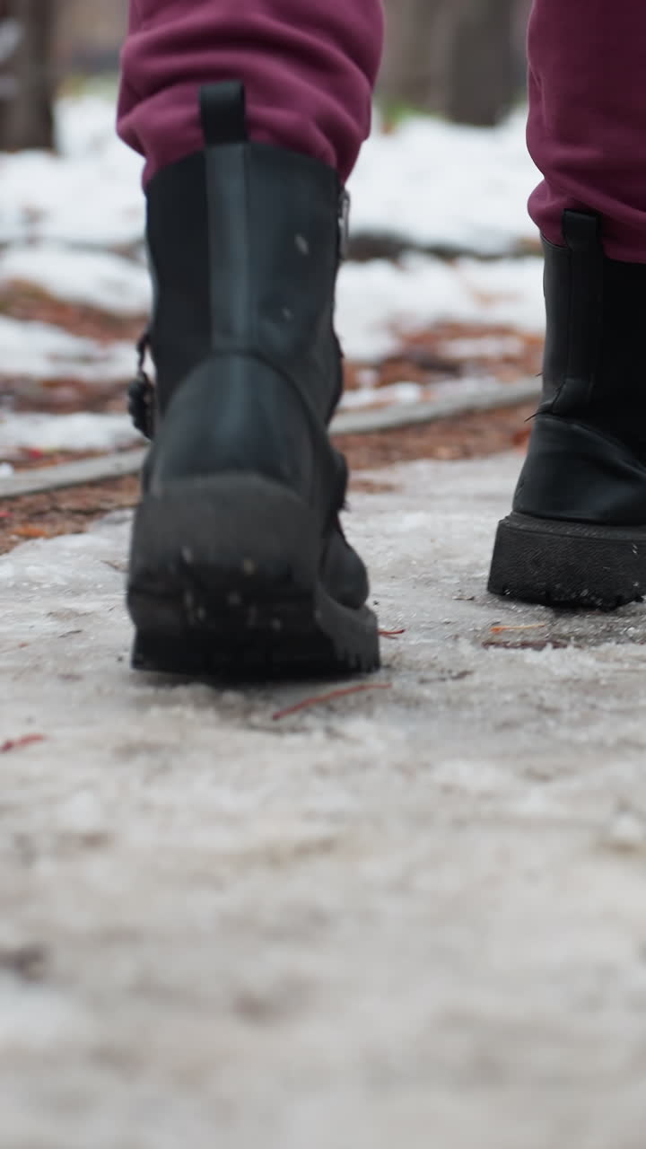vista trasera de cerca de una persona con botas negras pisando un camino cubierto de nieve helada esparcido de hojas secas, con un fondo borroso de árboles y figuras distantes