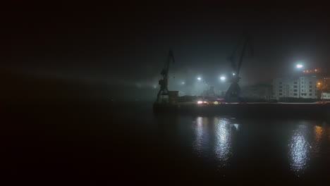 misty industrial harbor with silhouetted cranes and reflective water at night.