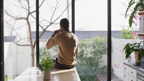 Happy african american man sitting at countertop in kitchen, drinking coffee